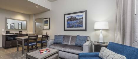 A modern hotel room with a grey sofa, a blue sofa, a dining area with a table and chairs, a small coffee table, a lamp, and a framed picture on the wall.