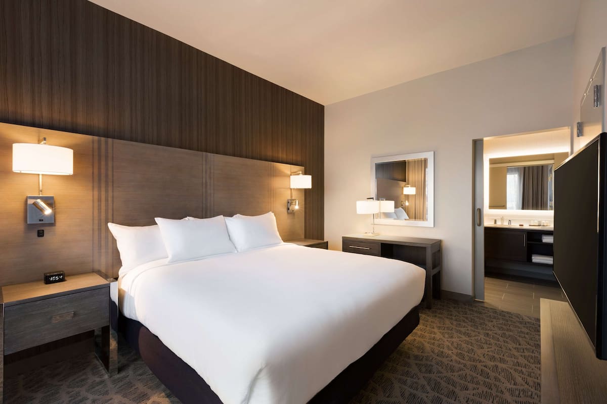A hotel room with a large bed, bedside tables, a mirror, a TV, and a bathroom visible through an open door.
