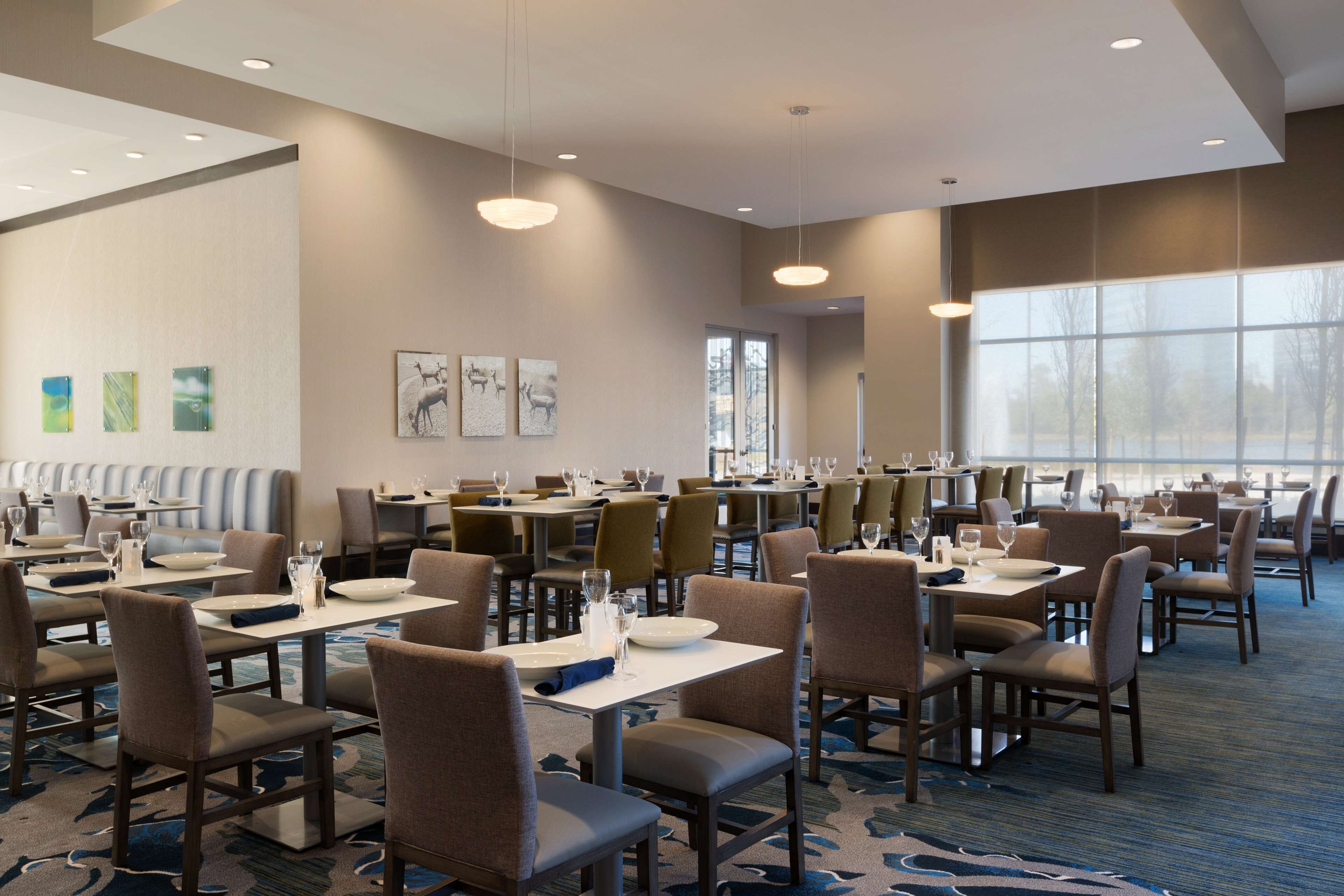 A spacious dining area with multiple tables and chairs, large windows providing natural light, and artwork on the walls.