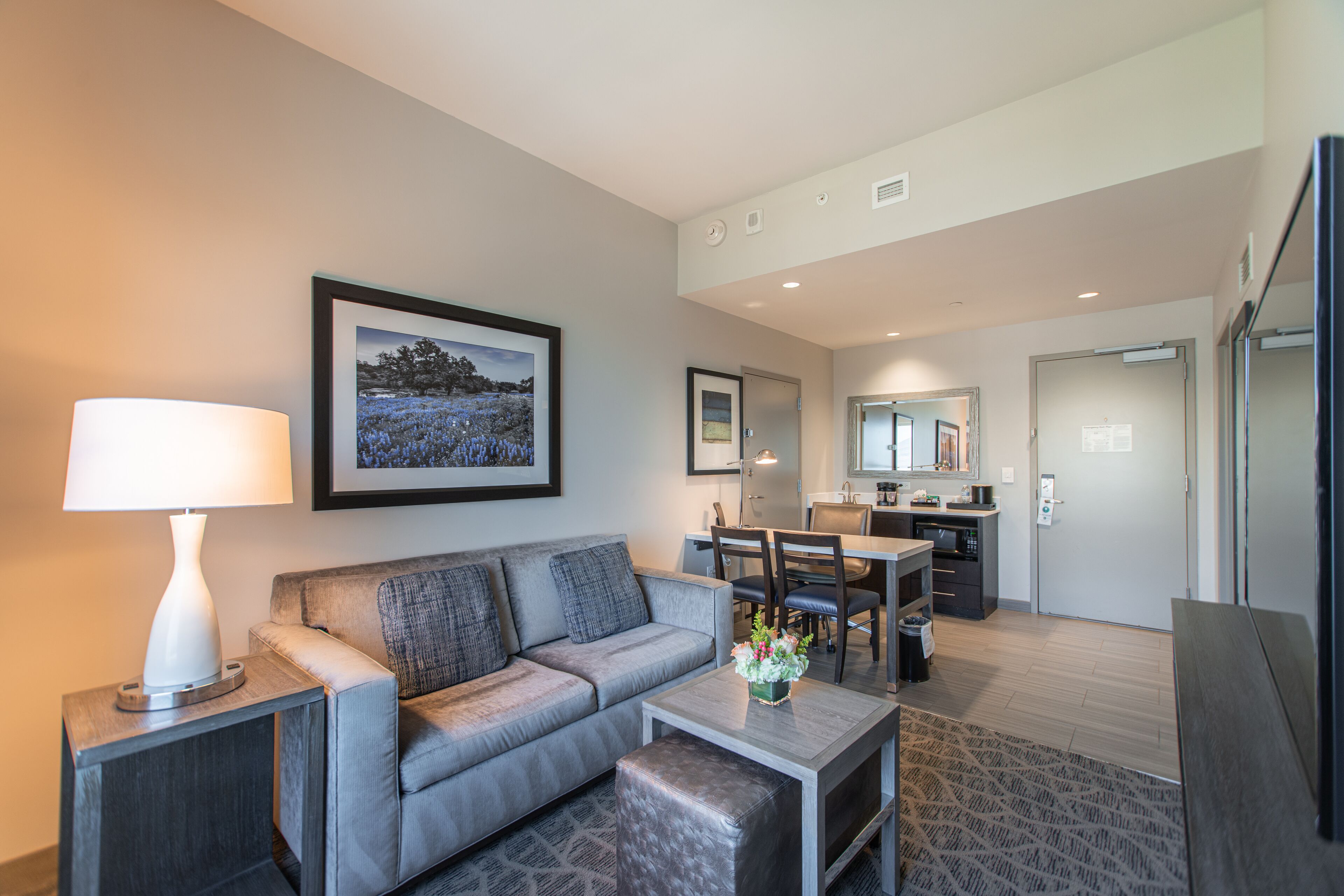 A modern hotel room with a grey sofa, a small table with a lamp, a framed picture on the wall, a dining area with chairs, and a kitchen area.