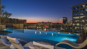 Una piscina en la azotea con sillones de descanso, el horizonte de la ciudad al fondo y un atardecer.