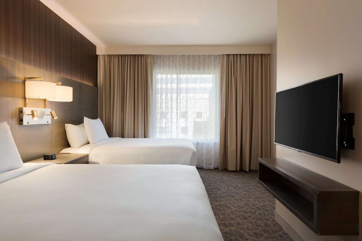 A hotel room with two beds, a TV, a wall-mounted lamp, and a window with curtains.