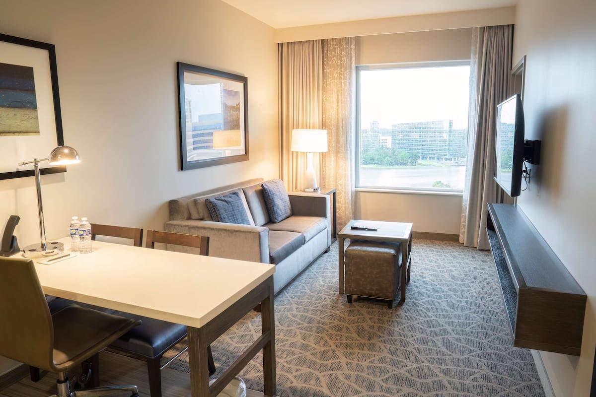 A hotel room with a sofa, a dining table, a TV, and a window with a view of buildings.