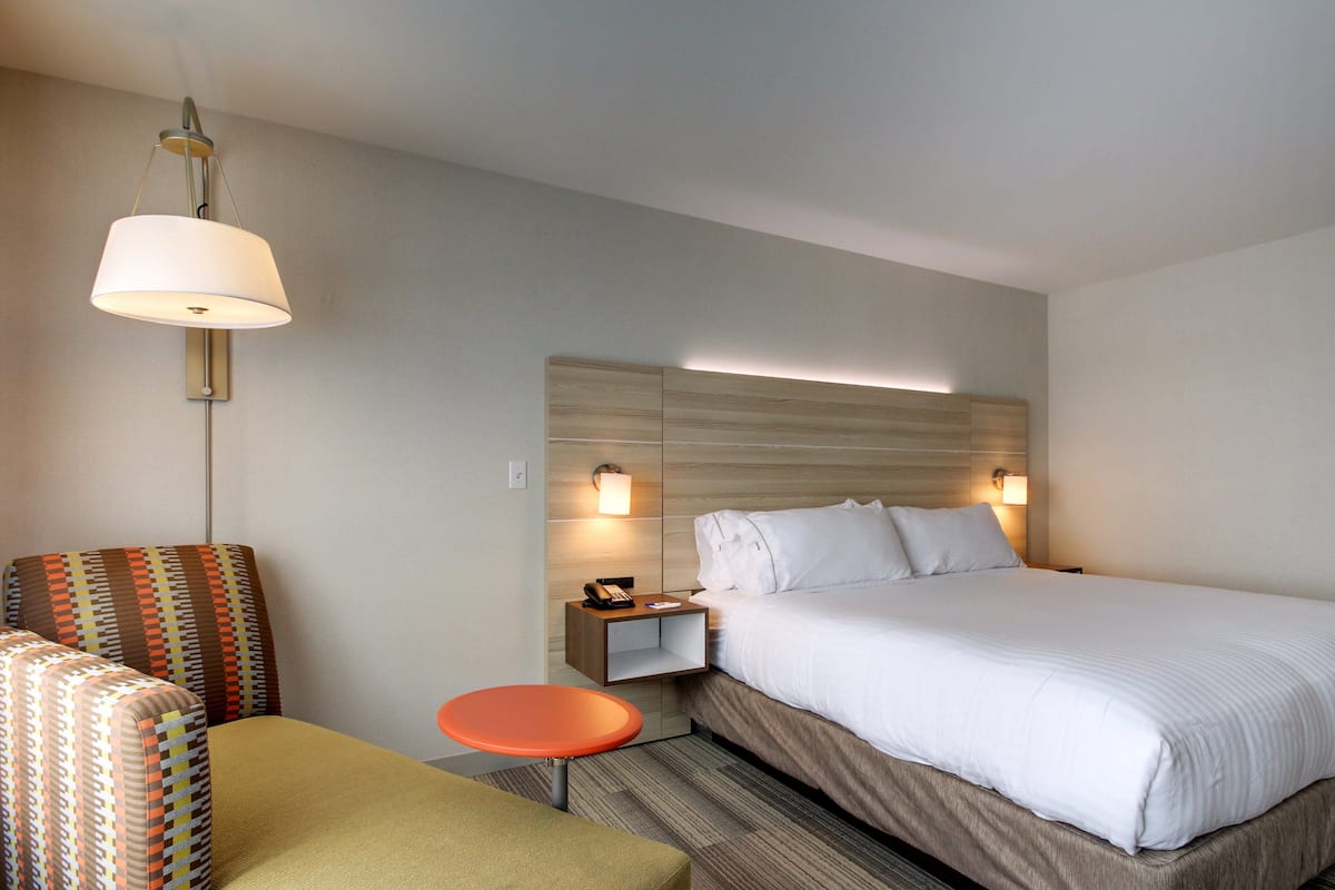 A hotel room with a large bed, a nightstand with a lamp, a small table, and two patterned armchairs.