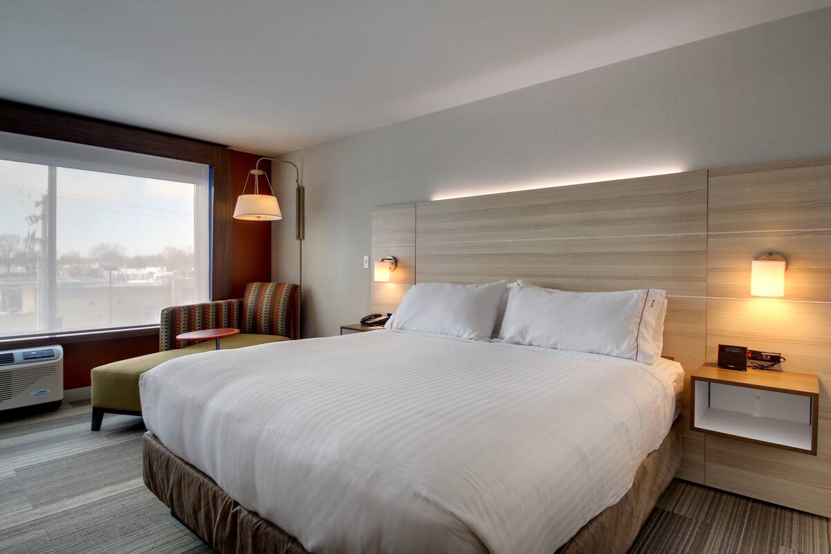 A hotel room with a large bed, a chair, a nightstand, and a window with a view of the city.