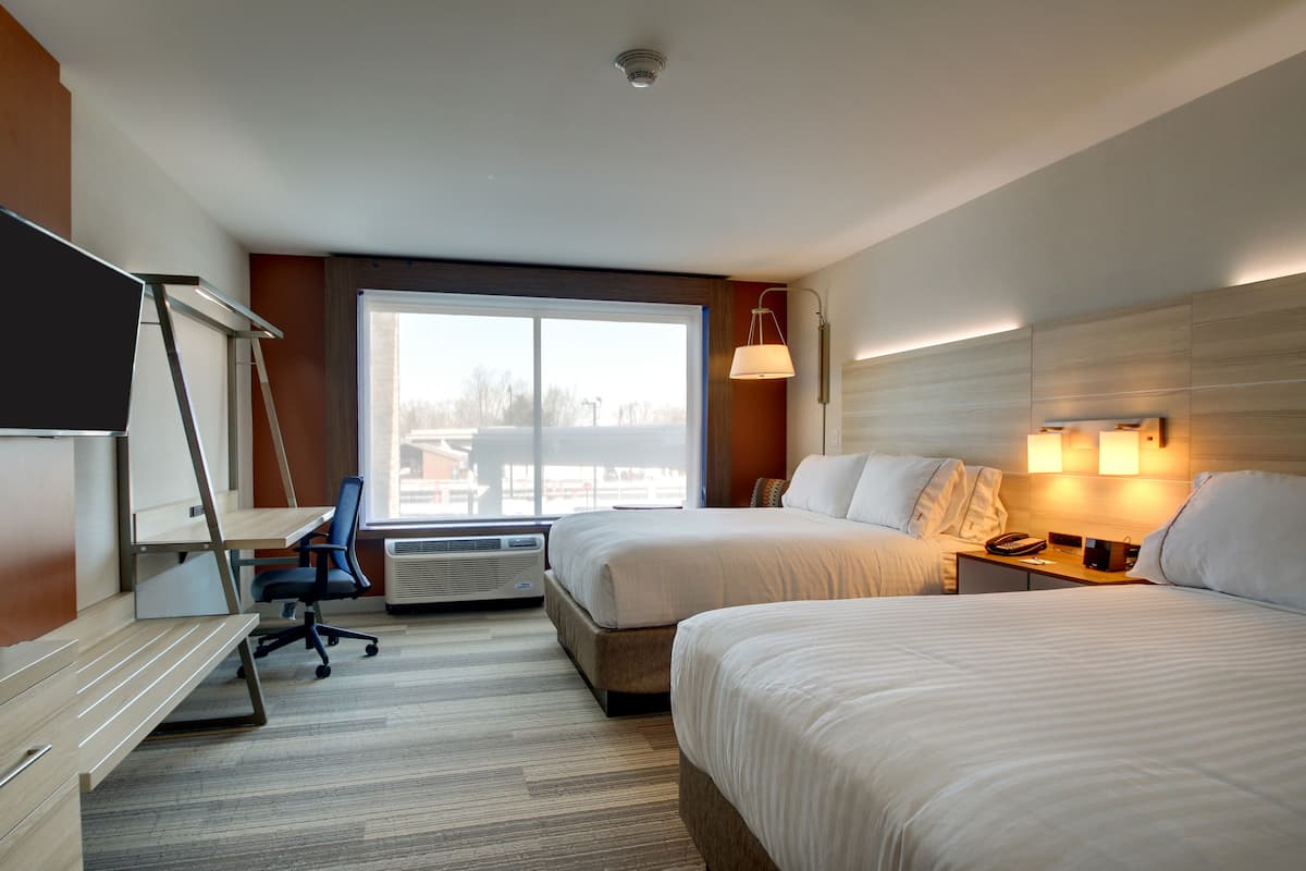 A hotel room with two beds, a desk, and a chair. There is a large window providing a view of the outside.