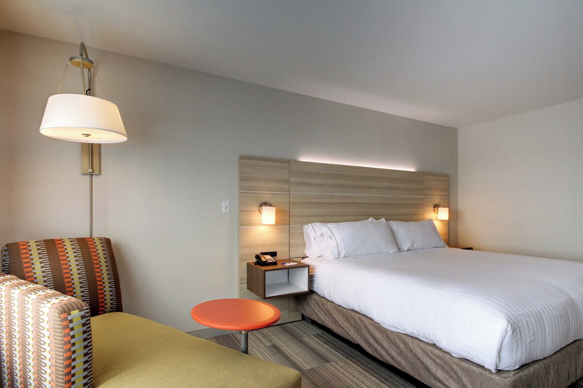 A hotel room with a large bed, a nightstand with a lamp, a small table, and two patterned armchairs.
