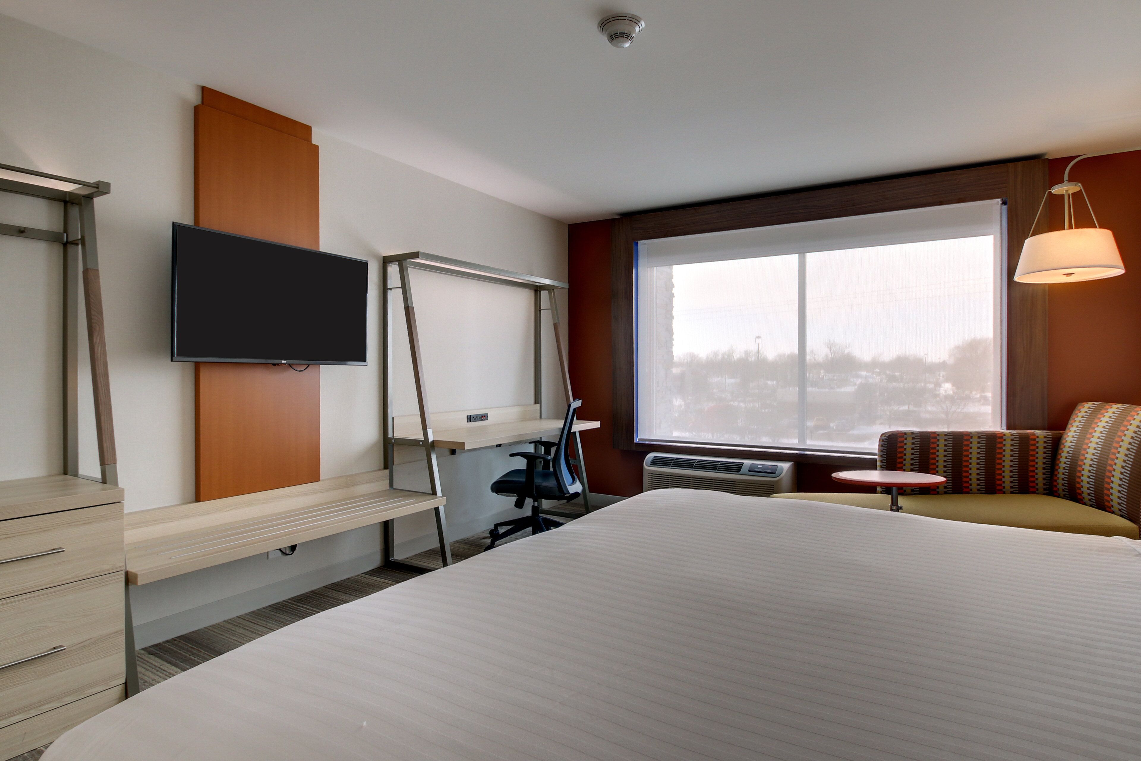 A hotel room with a large bed, a desk with a chair, a television, a window with a view, and a small table with a lamp.