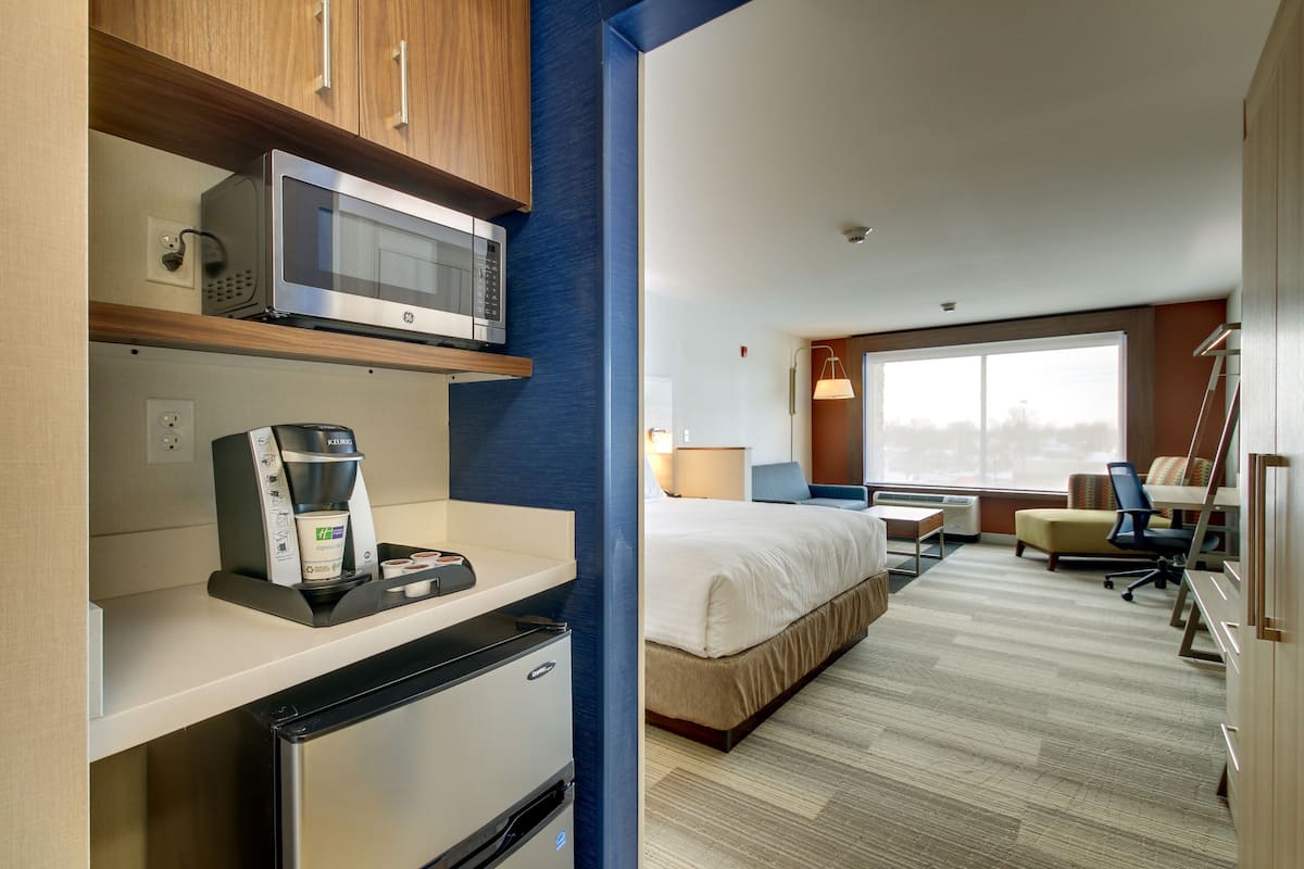A hotel room with a kitchenette, a bed, a desk, and a large window.