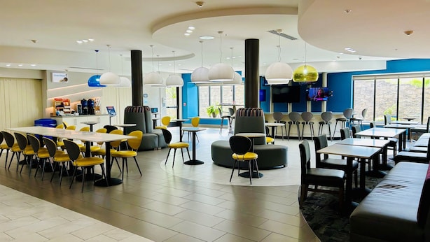 Un espace repas moderne avec des tables rondes et des chaises jaunes, une grande fenêtre offrant une belle vue et un téléviseur à écran plat fixé au mur.