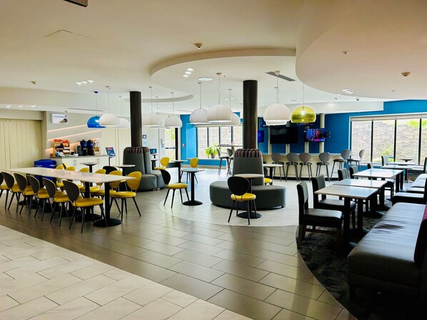 A modern dining area with round tables and yellow chairs, a large window with a view, and a flat-screen TV mounted on the wall.