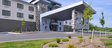 A modern hotel with a flat roof, multiple windows, and a landscaped front yard with small shrubs and a tree.