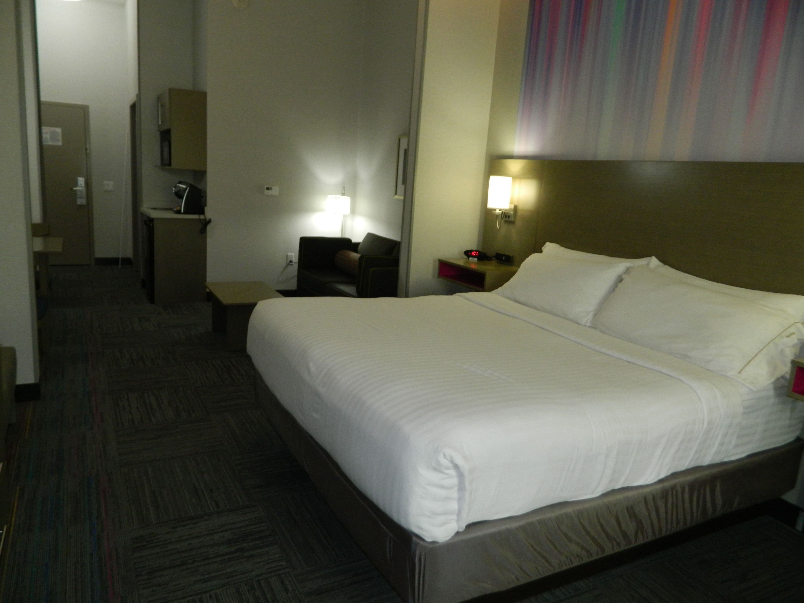 A hotel room with a large bed, a desk, a chair, a nightstand, and a wall-mounted lamp.