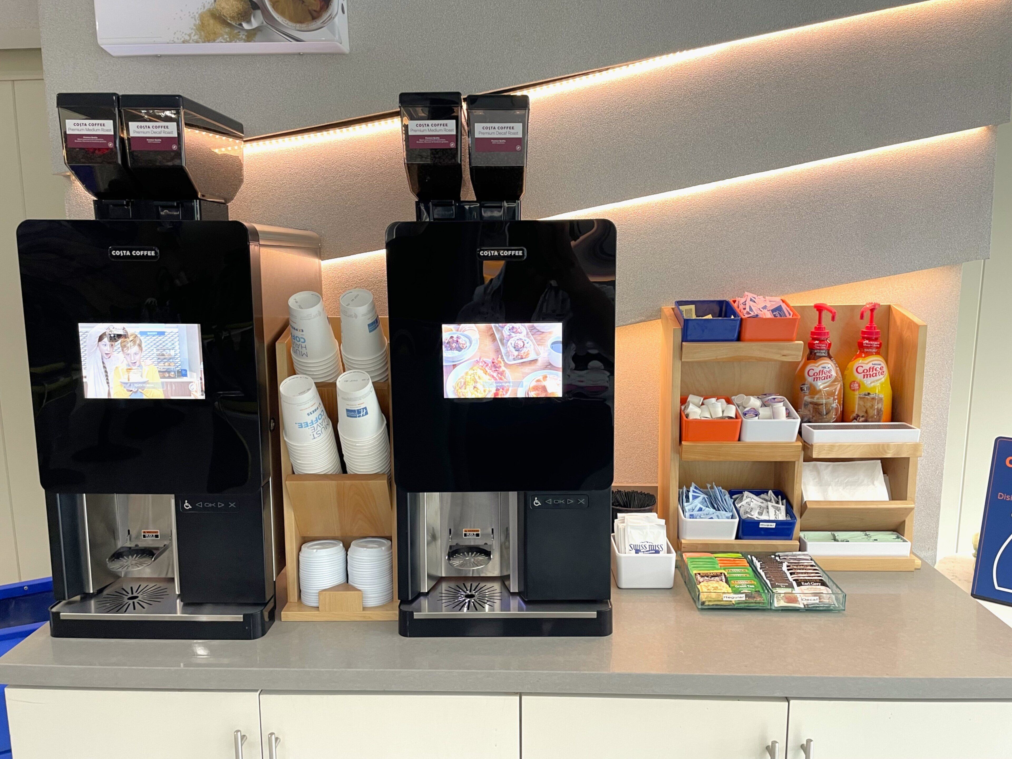 Three coffee machines with disposable cups, a coffee dispenser, and a shelf with coffee-related items.