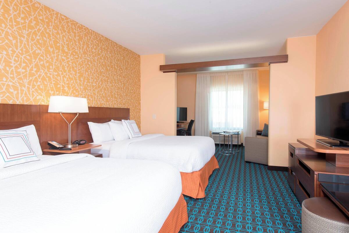 Quarto de hotel com duas camas, uma escrivaninha, televisão e banheiro visível através de uma porta aberta.