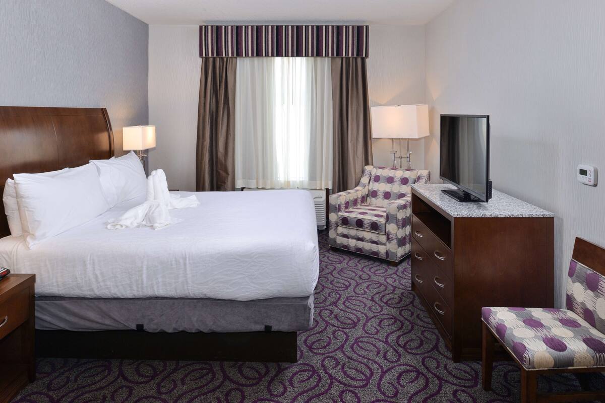 A hotel room with a large bed, a desk with a TV, a chair, and a lamp.