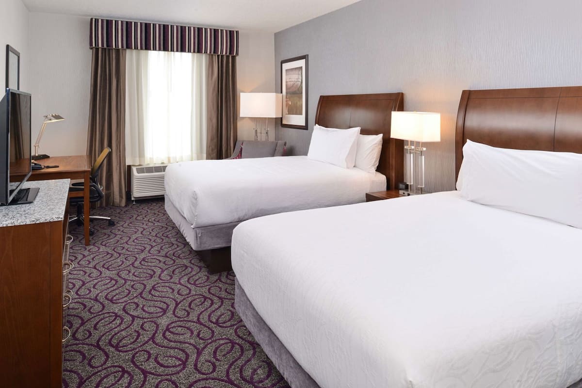 A hotel room with two beds, a desk, a TV, and a window with curtains.