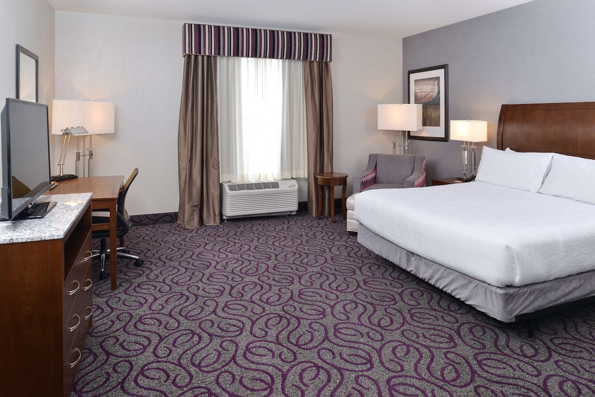 A hotel room with a large bed, a desk with a computer, a chair, a small table, and a wall-mounted TV.