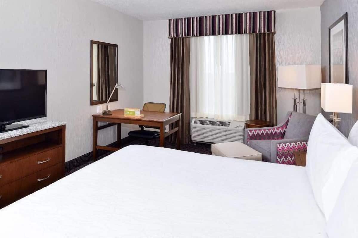 A hotel room with a bed, a desk, a television, a chair, a lamp, and a window with curtains.