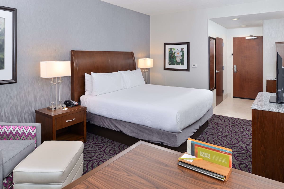 A hotel room with a large bed, a desk with a lamp, a chair, and a small table with a book.