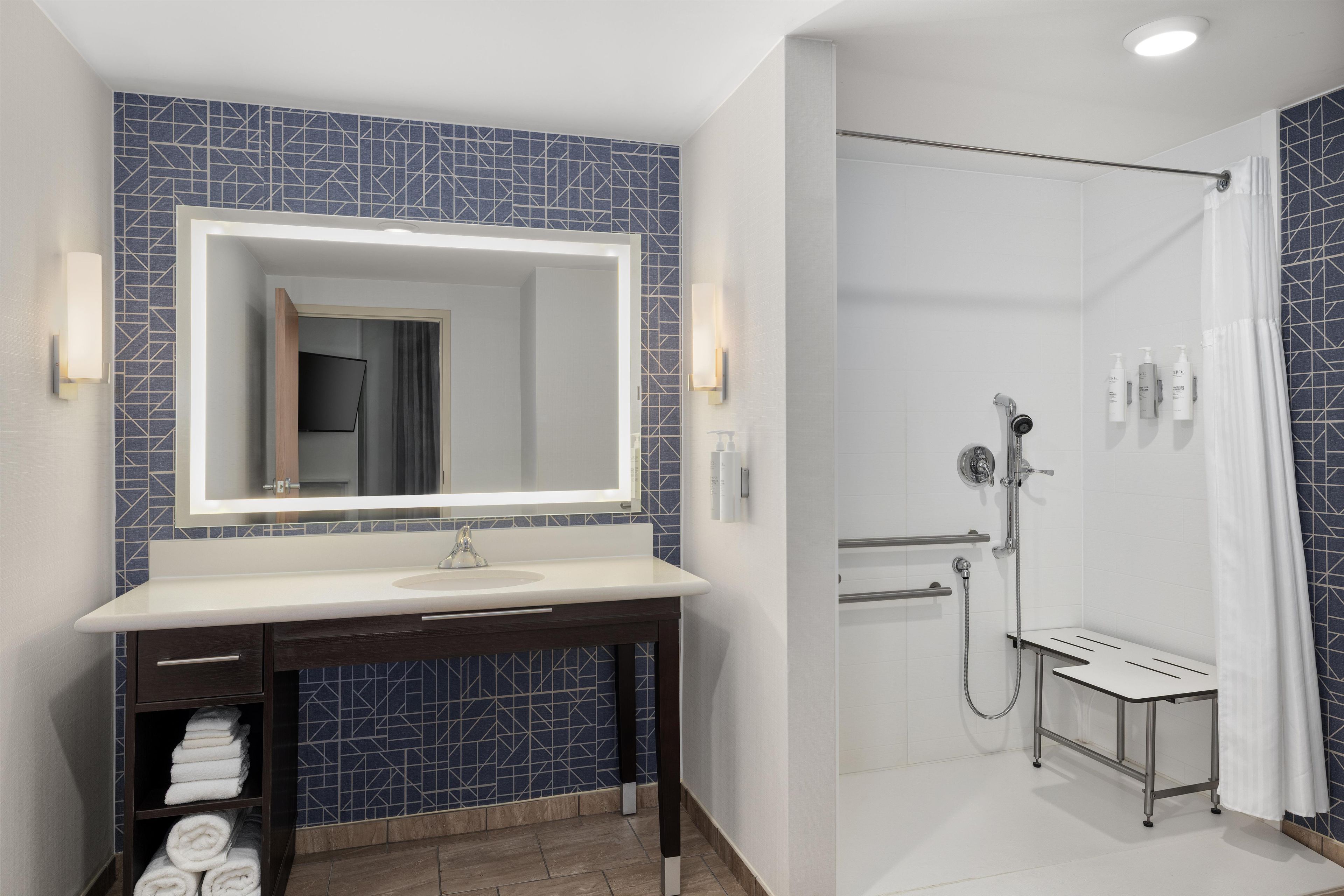 Un baño moderno con un espejo grande, un lavabo, una ducha con cabezal de ducha manual y un perchero para toallas.