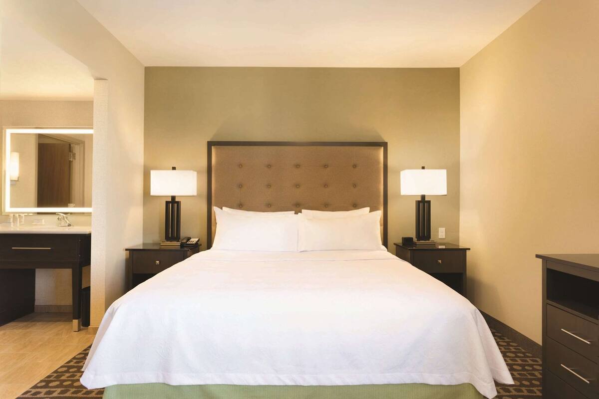 A hotel room with a large bed, two bedside tables with lamps, a nightstand, a dresser, and a mirror.