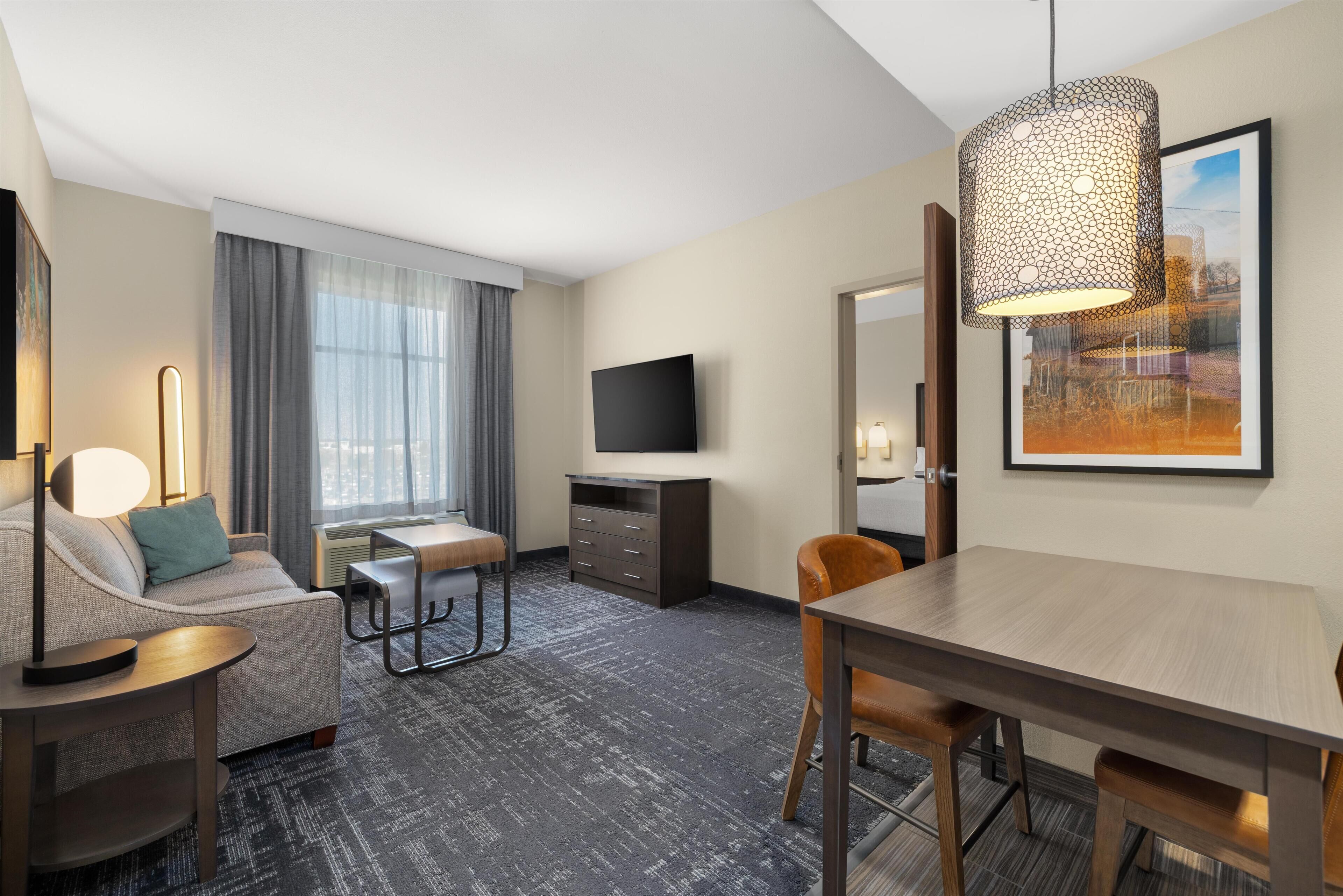 A hotel room with a sofa, a dining table, a TV, and a large window with curtains.