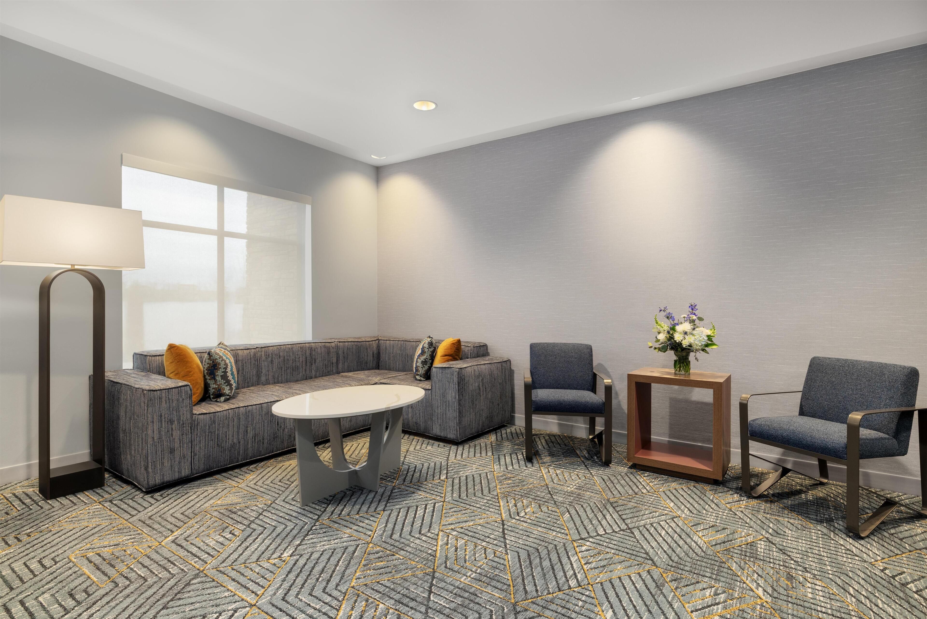 A modern hotel room with a sectional sofa, two armchairs, a small table, and a lamp.