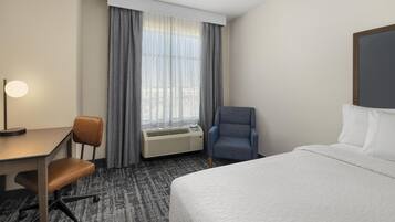 A hotel room with a bed, a desk with a chair, a standing chair, a window with curtains, and a wall-mounted air conditioner.