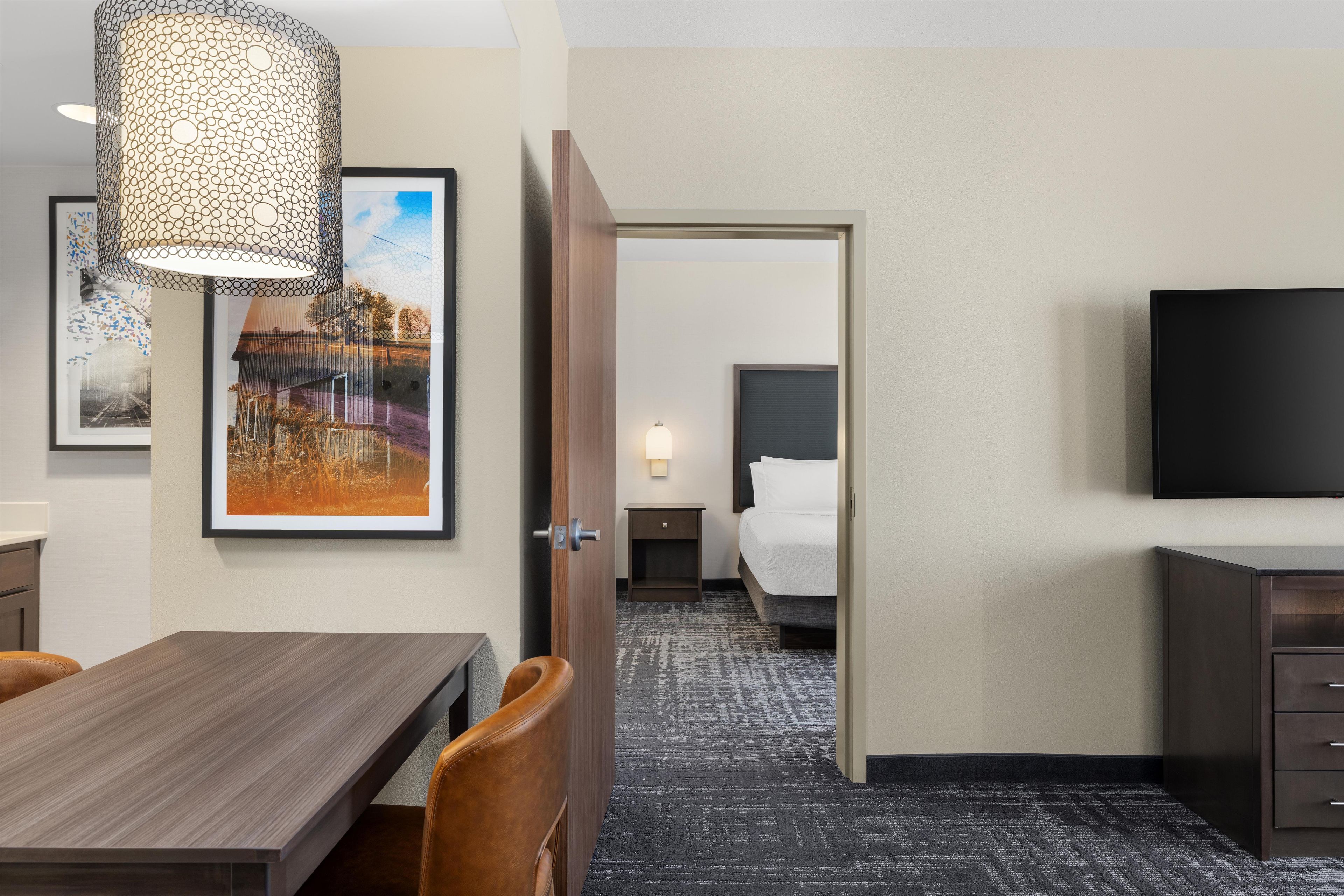 A hotel room with a bed, bedside table, lamp, TV, and a dining area with a table and chairs.