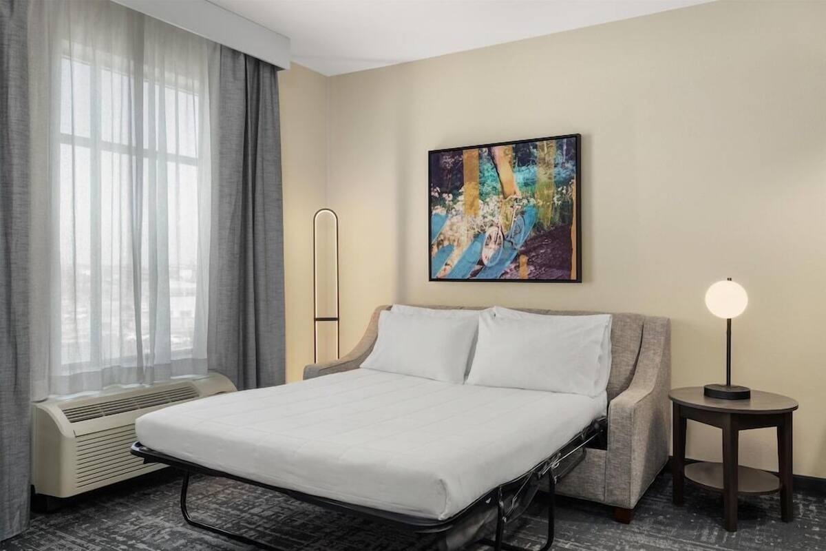 A hotel room with a bed, a chair, a table, a painting, and a window with curtains.