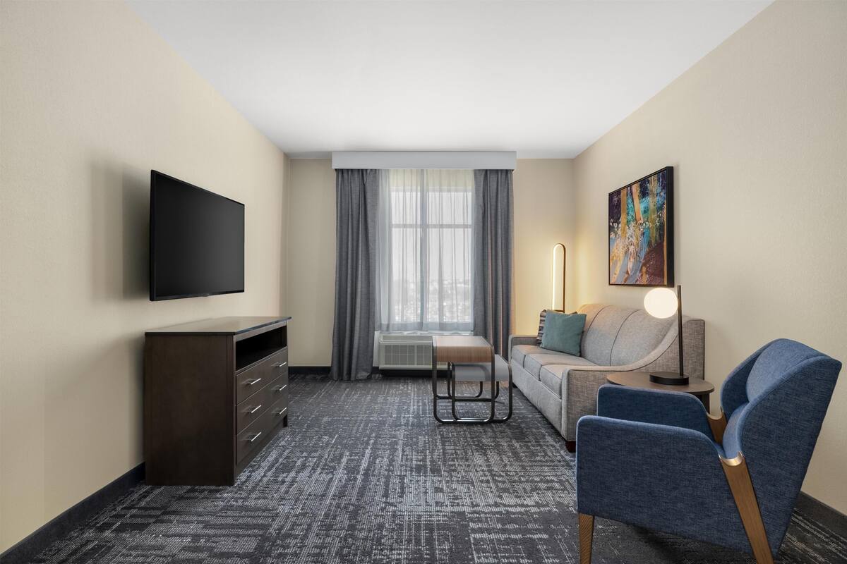 Habitación de hotel con una televisión de pantalla plana, un sofá, una silla, un escritorio, una lámpara y un cuadro en la pared.