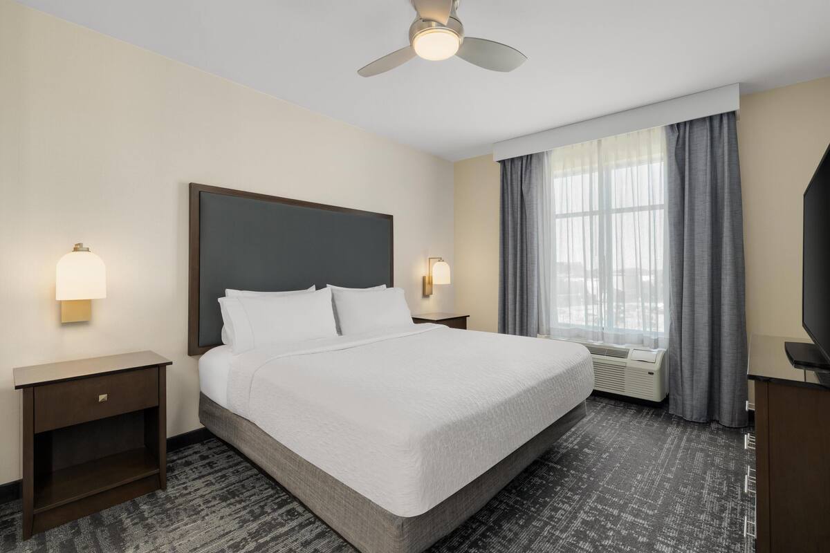 Habitación de hotel con cama, mesitas de noche, ventilador de techo, ventana con cortinas y televisor.