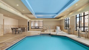 Una piscina interior con borde de azulejos, techo con iluminación empotrada y vista a un edificio a través de grandes ventanales.
