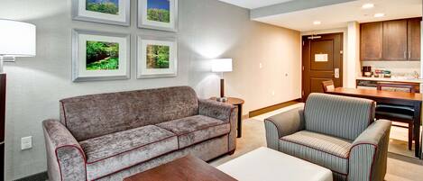 A modern living room with a sofa, armchair, and coffee table, a kitchen area with cabinets and appliances, and framed pictures on the wall.