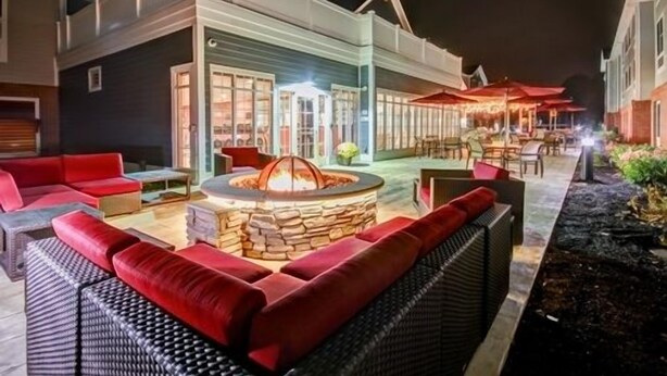 An outdoor seating area with a stone fire pit, red sofas, and a dining area with umbrellas. - Homewood Suites by Hilton Bridgewater/Branchburg (Branchburg)