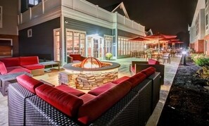 An outdoor seating area with a stone fire pit, red sofas, and a dining area with umbrellas. - Homewood Suites by Hilton Bridgewater/Branchburg (Branchburg)