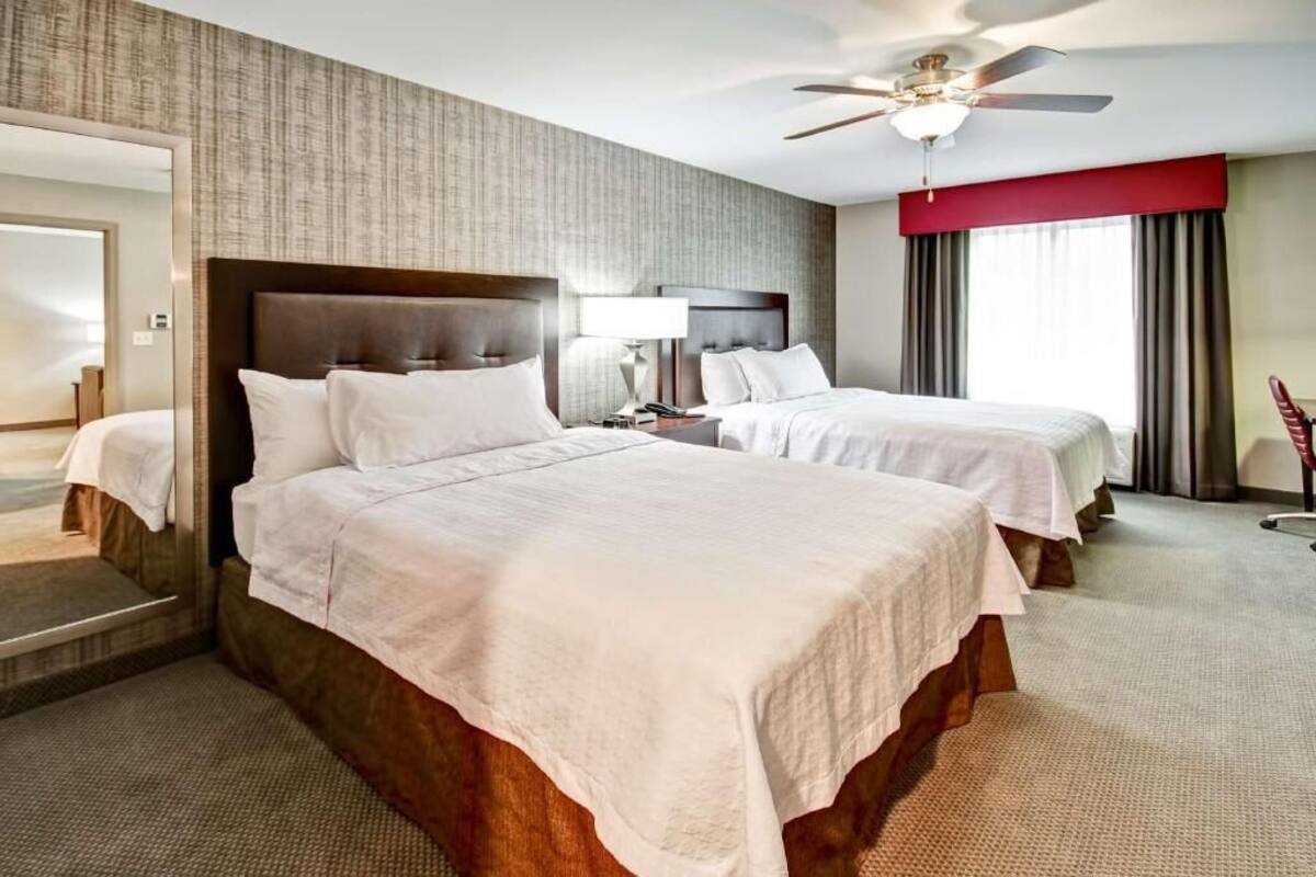 A hotel room with two beds, a ceiling fan, a large mirror, and a view of the outdoors.