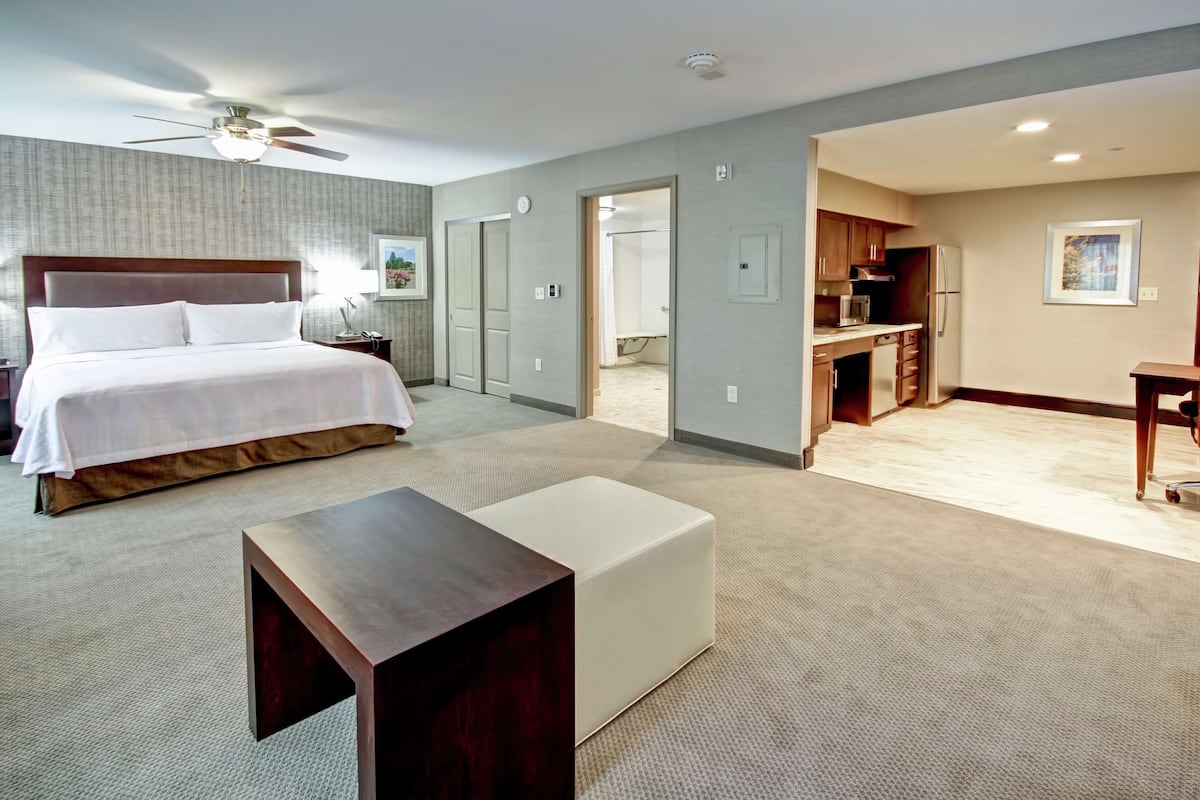 A bedroom with a bed, a desk, a chair, a ceiling fan, and a kitchenette.