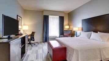 A hotel room with a large bed, a desk with a TV, a chair, a bench, and a window with curtains.