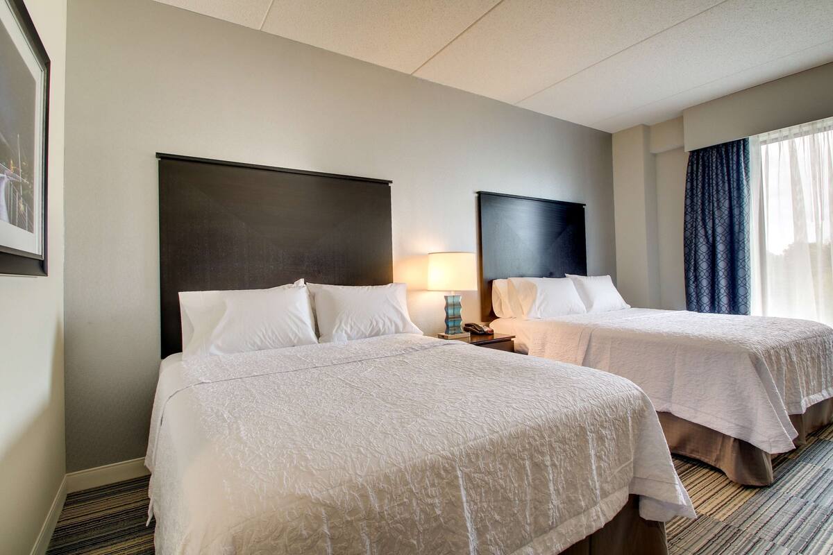 Habitación de hotel con dos camas, una mesita de noche con lámpara y una ventana con cortinas.
