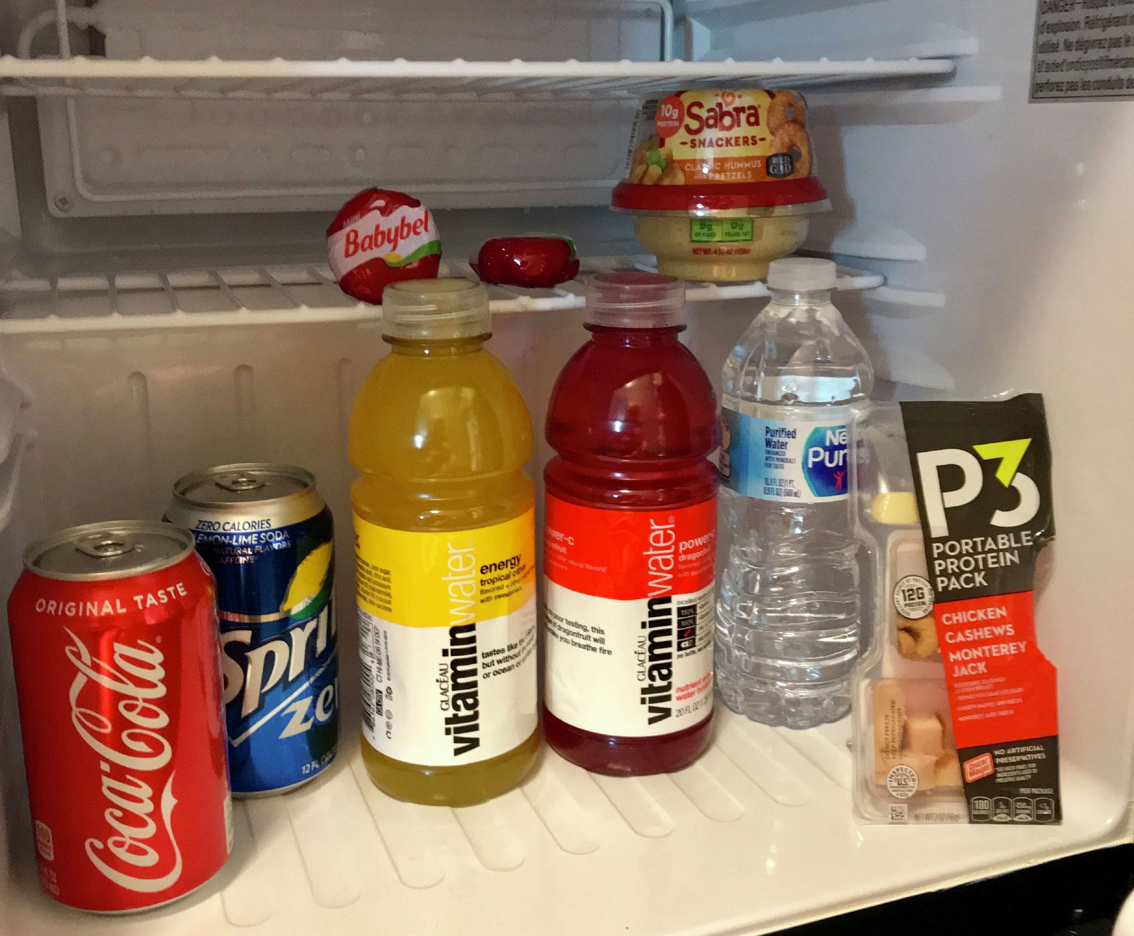 Uma geladeira com diversos alimentos e bebidas, incluindo uma lata de Coca-Cola, uma garrafa de Vitamin Water, uma garrafa de Gatorade, uma garrafa de Powerade, uma garrafa de água, um pote de comida para bebês, um pote de proteína em pó e um pote de lanches.