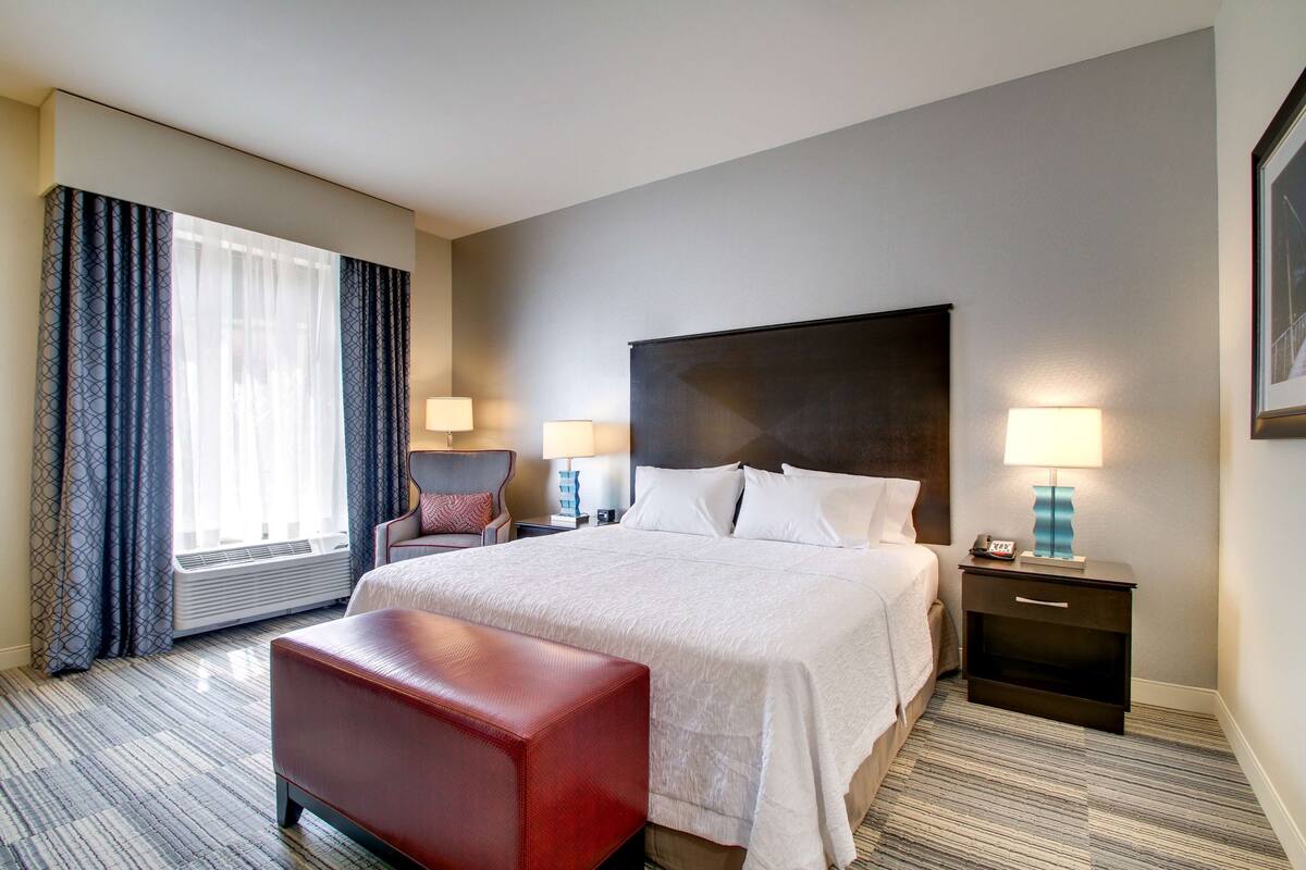 Una habitación de hotel con una cama grande, un banco rojo, dos mesitas de noche con lámparas, una silla y una ventana con cortinas.