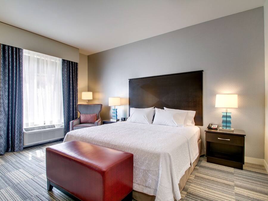 A hotel room with a large bed, a red bench, two bedside tables with lamps, a chair, and a window with curtains.