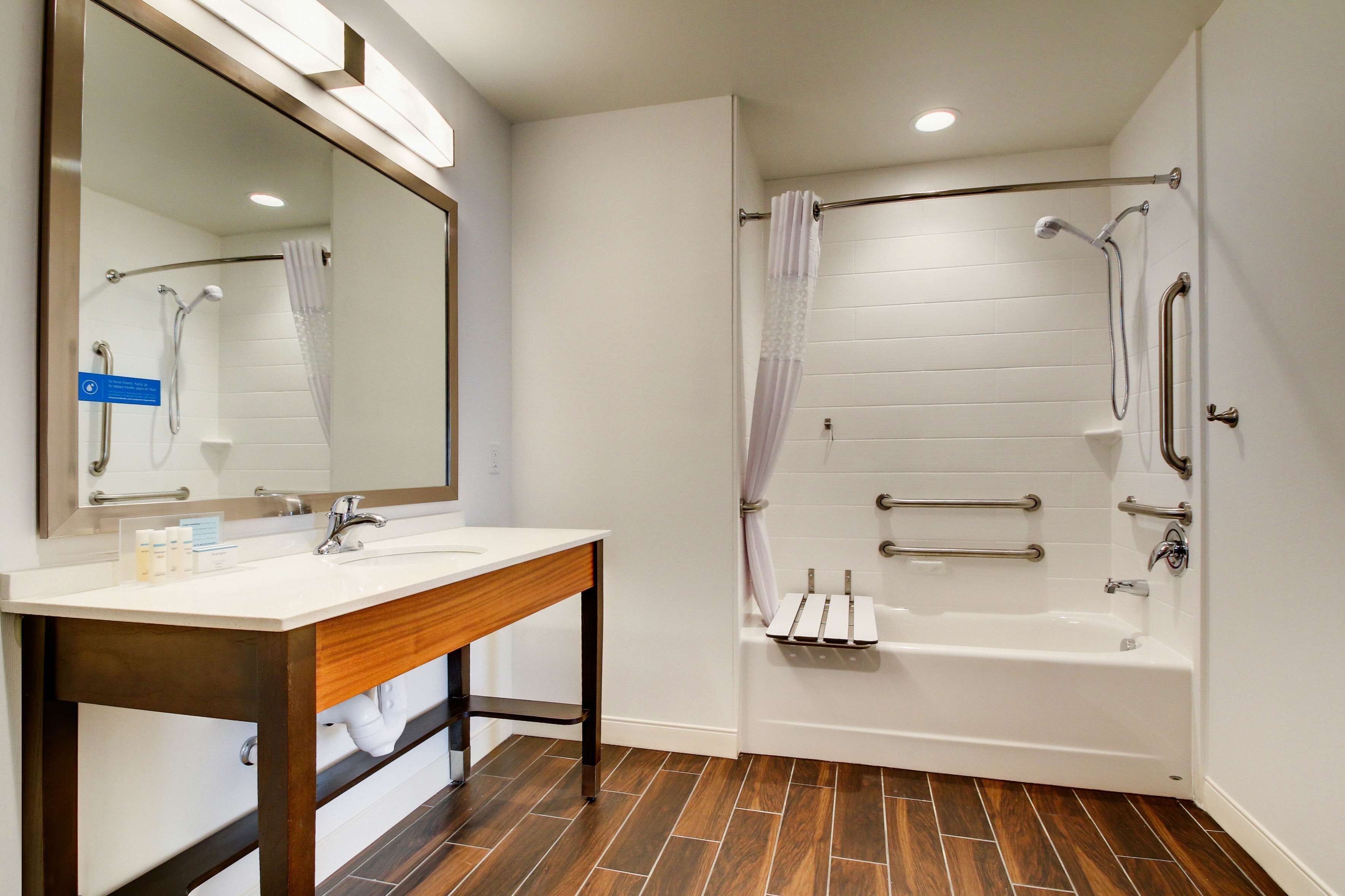 A modern bathroom with a wooden vanity, a large mirror, a bathtub with grab bars, and a shower area.