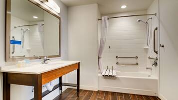 A modern bathroom with a wooden vanity, a large mirror, a bathtub with grab bars, and a shower area.
