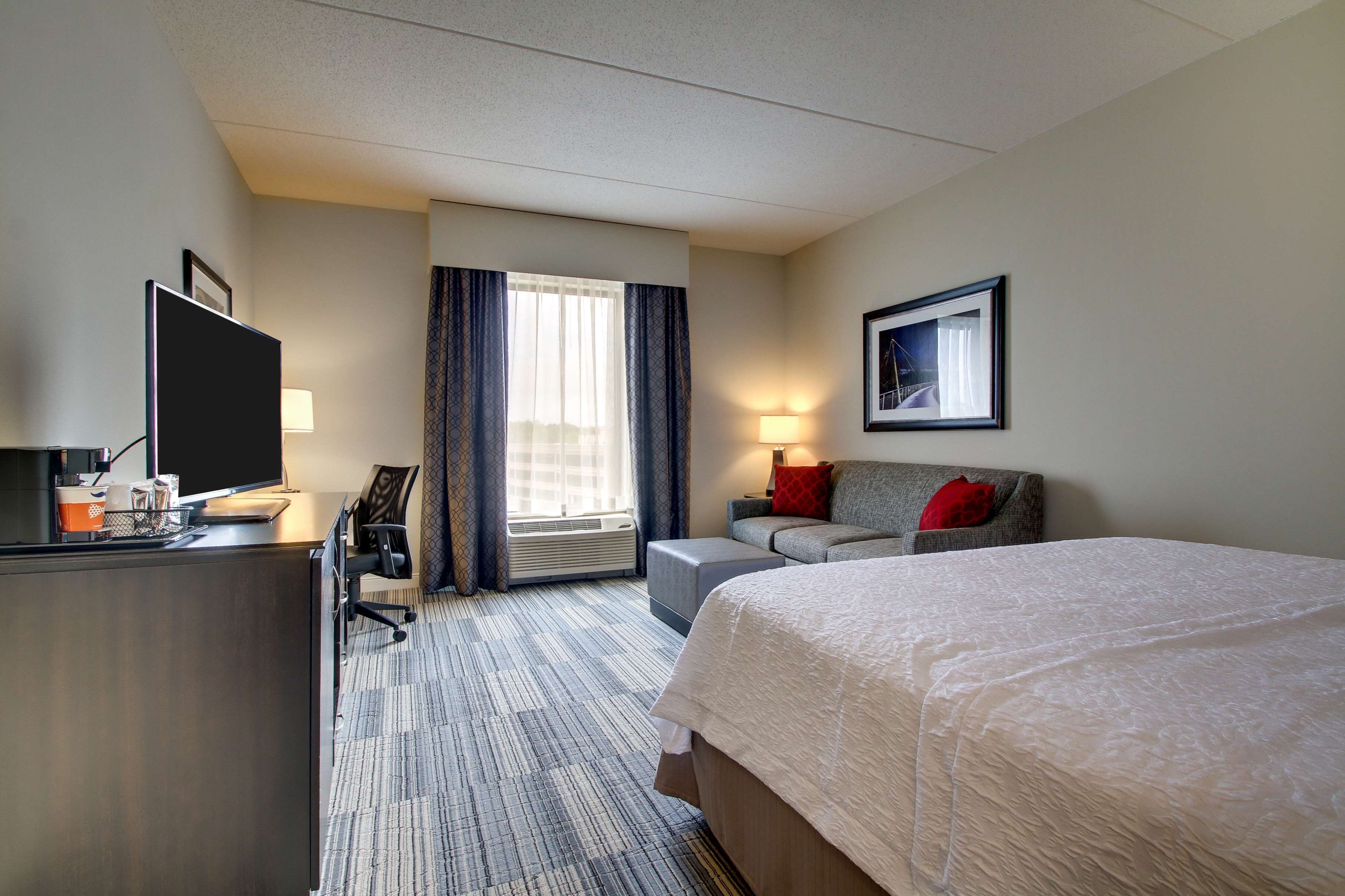 A hotel room with a large bed, a desk with a computer, a chair, a sofa, and a window with curtains.