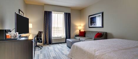 A hotel room with a large bed, a desk with a computer, a chair, a sofa, and a window with curtains.