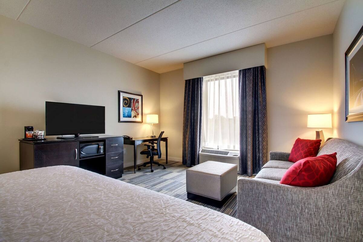Habitación de hotel con cama, escritorio, silla, televisión y ventana con cortinas.