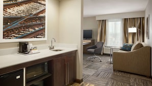 A hotel room with a kitchenette, a sofa, a TV, and a large window with curtains. - Hampton Inn & Suites Minooka (Minooka)